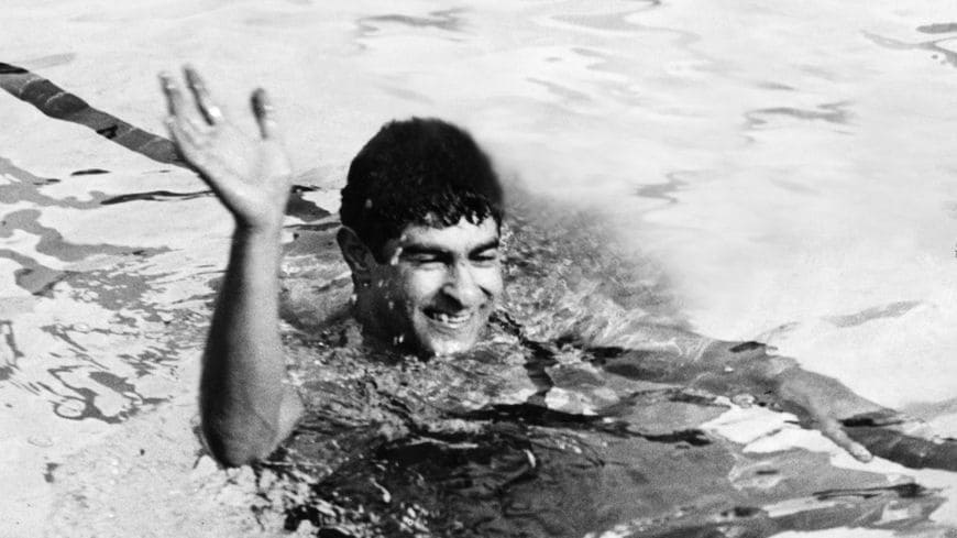 Nakache faisant un grand signe de la main, tout sourire, dans une piscine.